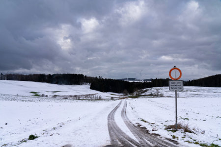 A winter landscape features a snow-covered road meandering towards a distant forest. The sky is filled with dark clouds, hinting at changing weather.の写真素材