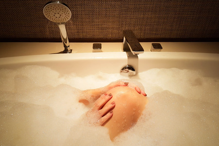 Woman in bathtub with fully foambath cleaning her leg with two hands in sexy mood; shoot by mobile camera in orange light and shadow style image.の写真素材