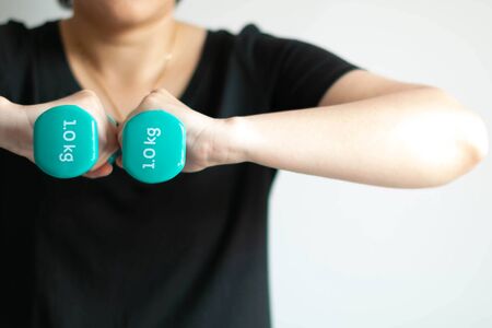Close-up of woman use two hands lifting pastel-blue dumbbells weighing 1 kilogram in each side; at center of chest on white background. health care and exercise concept.の写真素材
