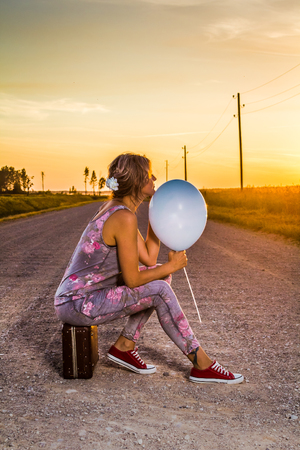 woman sitting on an old suitcase in the middle of a rural road, with a blue balloon in her handの写真素材