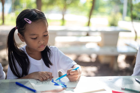 young girls are sitting at a table with markers and coloring books. They are drawing and coloring together, enjoying their time in the park. Concept of fun and creativityの写真素材