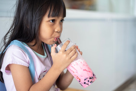 A young girl is drinking a pink beverage from a straw. The drink is in a clear cup, and the girl is wearing a pink shirt. The scene is casual and relaxed, with the girl enjoying her drinkの写真素材
