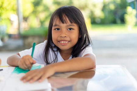 A young girl is sitting at a table with a pencil and a piece of paper. She is smiling and she is enjoying herself. The scene suggests a positive and happy atmosphereの写真素材