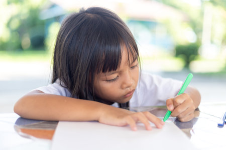 A young girl is sitting at a table writing with a pen. She is focused on her work and she is in a...