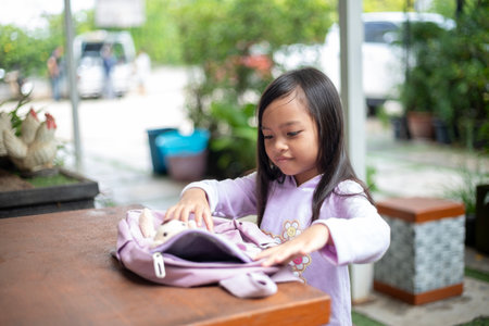 A young girl is sitting at a table with a backpack and a teddy bear. She is looking at the backpack and seems to be curious about what's insideの写真素材