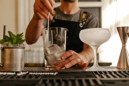 Detail of the hand of a waiter in an apron removing the ice from a glass with a container with a mental beside and a frost glass for cocktailの写真素材