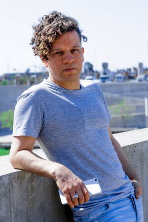 Vertical photo of a portrait of a man with curly hair holding a mobile phone while leaning against a wall with the city in the background out of focusの写真素材