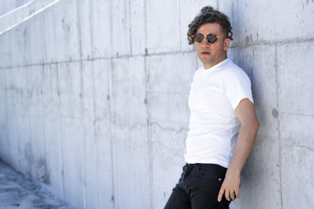 Stylish man with curly hair and sunglasses leaning against a concrete wall