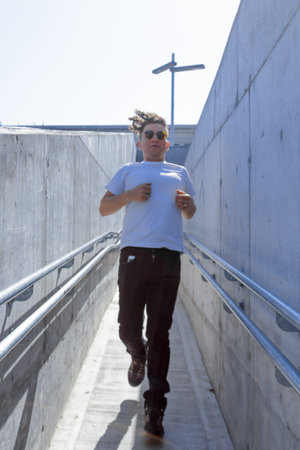 Vertical photo of a man with curly hair and sunglasses running down a narrow concrete passageway in...