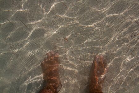 Feet of a person in the water covered by white sand and with the reflections of the sun on the waterの写真素材