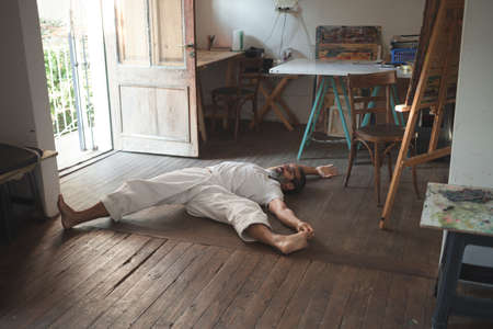Vertical photo of a man dressed in white with long, gray hair up and with glasses sitting stretching his back while he holds his foot with his hand doing a yoga pose in a wood studioの写真素材