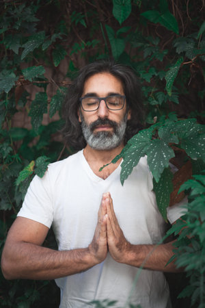 Vertical photo of the portrait of a man in white with long gray hair and glasses standing on a garden with his hands together in meditationの写真素材