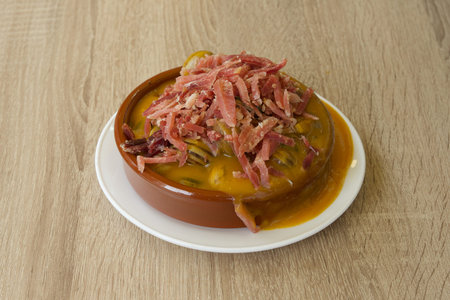 A plate of food with bacon and meat on top. The plate is on a wooden table. The food is served on a...