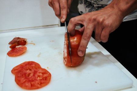 A person is cutting a tomato on a cutting board. The tomato is cut in half and the knife is in the...