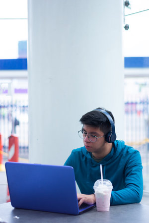 young Asian man working on laptop computer at modern location listening to music with headphones and drinking milkshakeの写真素材