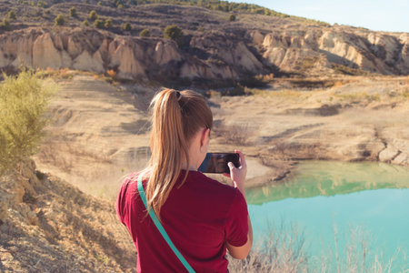 Young blonde female hiker with a ponytail wearing a red t-shirt and taking a photo of a blue lake in...
