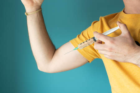 Powerful arm of a caucasian man wearing a yellow t-shirt on a turquoise background getting a vaccine with a syringeの写真素材