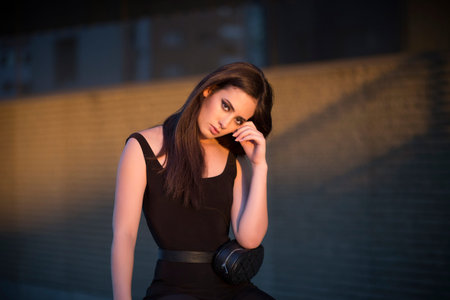 Attractive young female in black outfit looking at camera while standing near wall with hands behind...