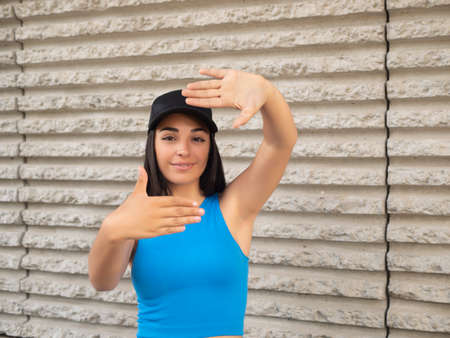 Friendly young Hispanic female in blue top and black cap showing photo gesture and looking at camera with smile against rough wall on city streetの写真素材