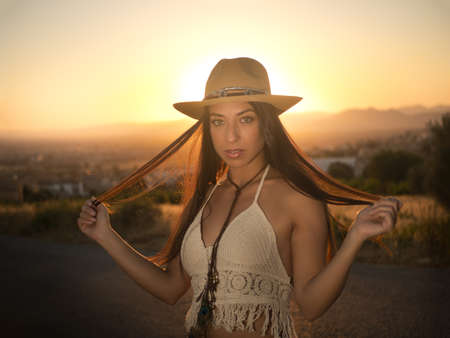 Calm brunette in crochet top and straw hat touching long hair and looking at camera while leaning on stone wall with metal net at sunsetの写真素材
