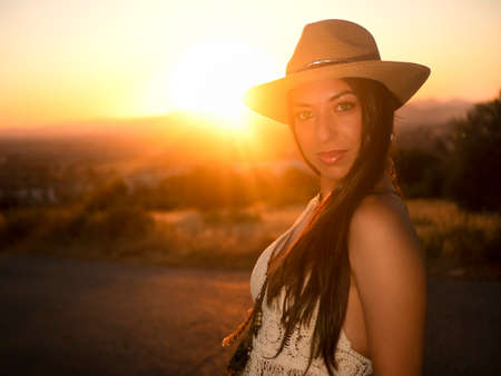 Calm brunette in crochet top and straw hat touching long hair and looking at camera while leaning on stone wall with metal net at sunsetの写真素材