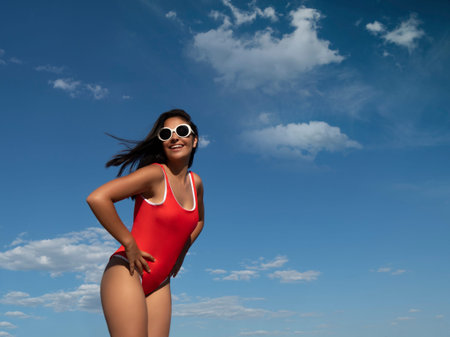 Charming shocked female in sunglasses and swimsuit standing with opened mouth against blue sky with...
