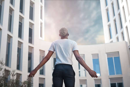 From below of African American male in elegant white shirt looking at camera while standing against modern skyscrapers in city on summer dayの写真素材