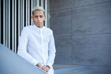 Serious African American male with dyed hair in sportswear looking at camera while sitting on street against blurred background on sunny summer dayの写真素材