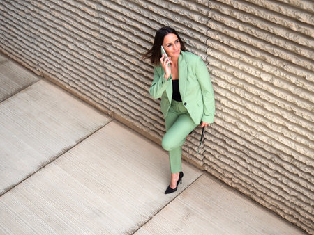 From above full body of content female entrepreneur looking up while speaking on cellphone near wall on street of cityの写真素材