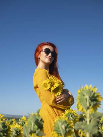 Delighted female with flying hair looking at camera with smile while holding yellow sunflower in field on summer day in countrysideの写真素材