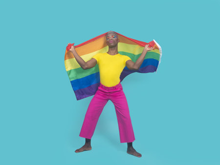 Full body happy African American man with LGBT flag wrapped around hips touching head and looking at camera with smile against turquoise backgroundの写真素材