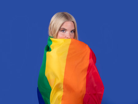 Happy young woman with blond hair wearing bright yellow bikini swimsuit standing under vivid rainbow umbrella on blue backgroundの写真素材