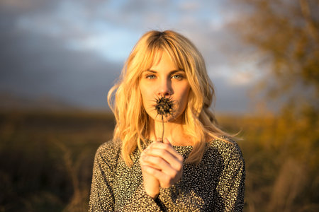 Graceful young calm female model with long blond hair in blouse with dry spiky plant in hand looking at camera in grassy meadow against cloudy sunset skyの写真素材