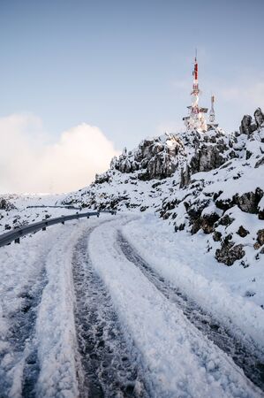 impressive background of a snowy mountain at sunset. Secondary road with tire tracks leading to a telecommunications tower surrounded by heavy snow in high mountainsの写真素材