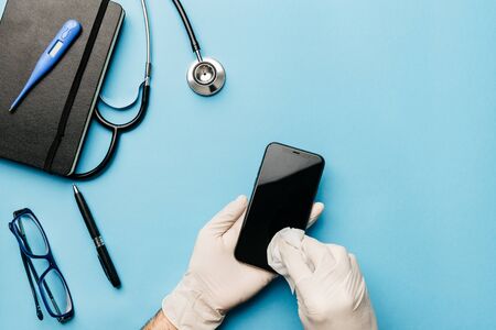 hands with gloves of a doctor desinfecting his smartphone after a practice in coronavirus epidemic in blue backgroundの写真素材