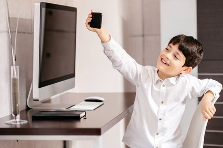 A boy standing in front of a computer taking a selfie with smartphoneの写真素材