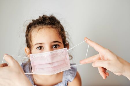 Girl with coronavirus mask prepared to go outside. Mothers hands putting the mask on her girl to prevent contagionの写真素材