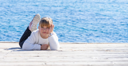 A young person reclines on a wooden pier, lost in thought, with the vast blue sea as their backdrop. The image captures a moment of youth and reflection, underscored by the infinite horizon and the tranquil maritime settingの写真素材