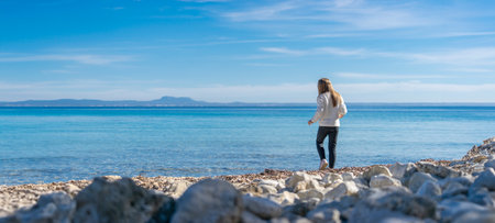 A child stands at the edge of a pebbled shore, gazing out towards the endless blue sea. The vastness of the ocean and the clear sky stretch out before them, inspiring wonder and the promise of discoveries beyond the horizonの写真素材