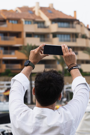 Rear view of an adult male taking a photo or selfie with a smartphone on vacation.