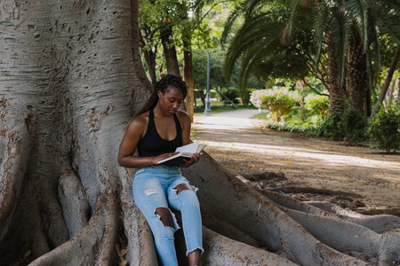 YOUNG AFRICAN AMERICAN WOMAN SITS RELAXING IN THE SHADE OF A TREE READING A BOOK
