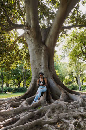 YOUNG AFRO-AMERICAN GIRL RELAXES READING WHILE SITTING IN THE SHADE OF A TREE.