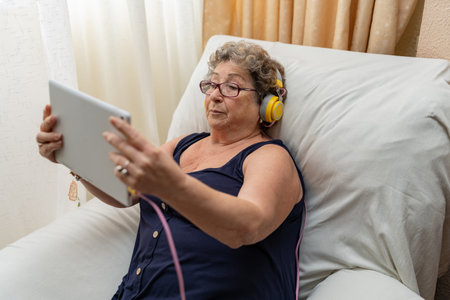 Older woman with headphones and glasses using a tablet while sitting on the sofa at home.の写真素材