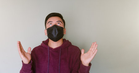 A Photo Young man wearing face mask looking up with praying hands and looking hopefulの写真素材