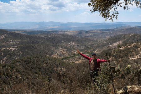 A man stands on a hill with his arms outstretched in the air, with a view of mountains in the distance.の写真素材