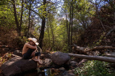 A man sits on a rock in a forest, wearing a cowboy hat.の写真素材
