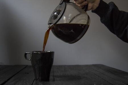 Glass jug pouring coffee into a black mug set on black wood and gray background.の写真素材