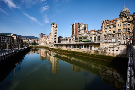 Cityscape of Bilbao shimmering in the Nervion river, featuring stunning reflections under a bright blue sky filled with cloudsの写真素材
