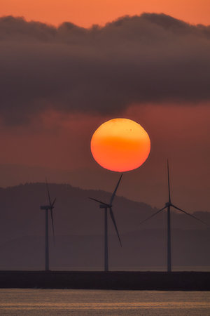 Wind turbines stand tall against the backdrop of a vibrant dawn, symbolizing the transition to...