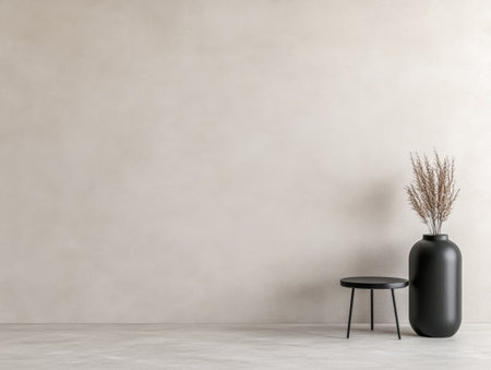 A stylish minimalist interior features a simple black stool beside a tall black vase filled with dried flowers against a soft, neutral wall and floor.の素材